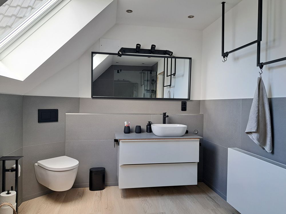 Badezimmer Ansicht Waschtisch: Bodenfliesen in Holzoptik, Wandfliesen in Grau. Waschtisch mit zwei Schubladen un Weiß, links daneben Toilette in Weiss. Armaturen, Kleiderstange über Heizung, Seifenspender und Lichtschalter in matt schwarz.