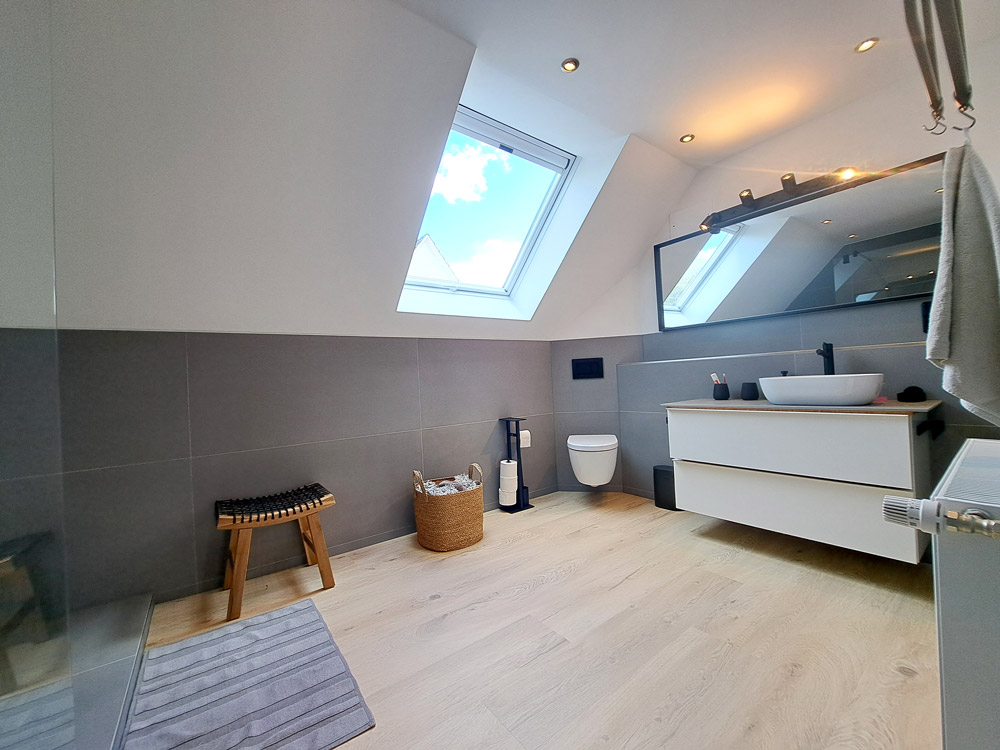 Badezimmer Ansicht Toilette: Bodenfliesen in Holzoptik, Wandfliesen in Grau. Waschtisch mit zwei Schubladen in Weiß, links daneben Toilette in Weiss, daneben Fenster. Armaturen, Seifenspender und Lichtschalter in matt schwarz.