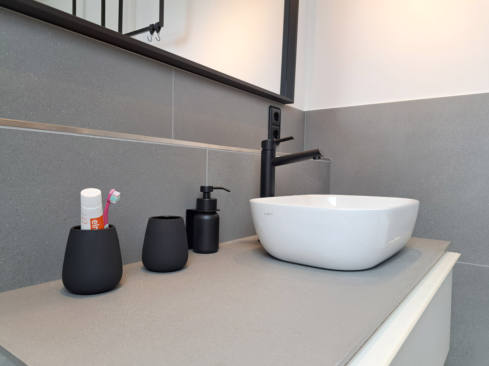 Badezimmer Detailansicht Waschbecken: Armaturen, Spiegelumrahmung, Seifenspenderm, Steckdosen und Becher in matt schwarz.