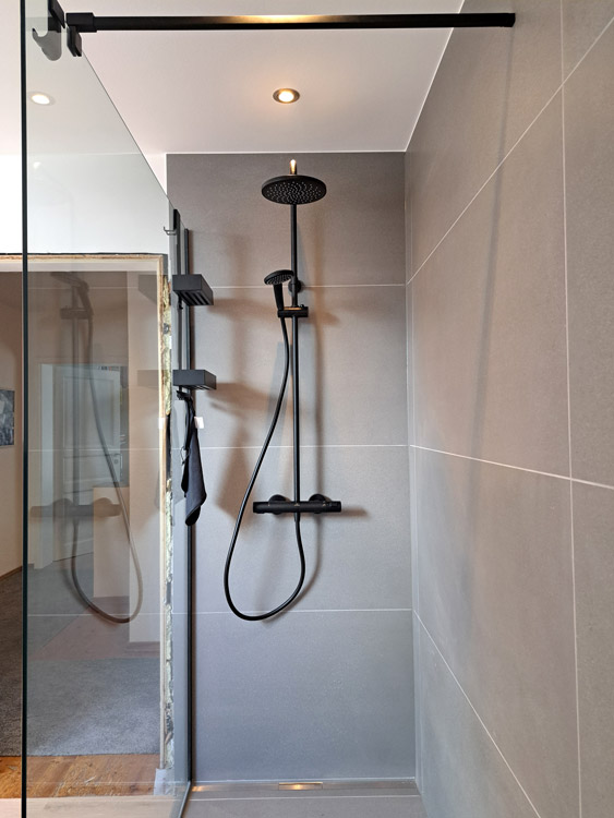 Dteailansicht Dusche mit Glaswand, Armaturen und Duschregal in Mattschwarz. Separater Duschkopf und Regendusche,
