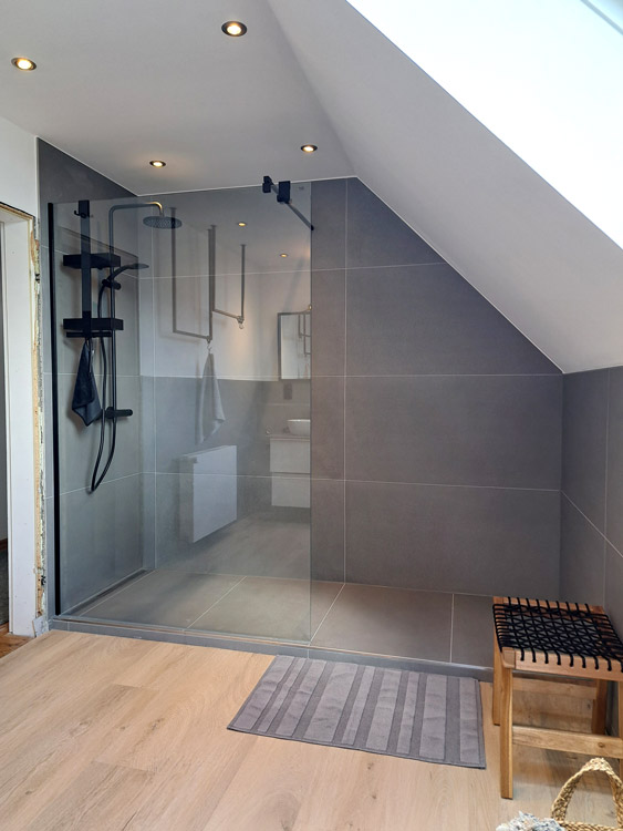 Dusche mit Glaswand, Fliesen in Hellgrau, Armaturen und Duschregal in Mattschwarz. Separater Duschkopf und Regendusche. Davor Hocker aus Holz und Badezimmerteppich in Grau.